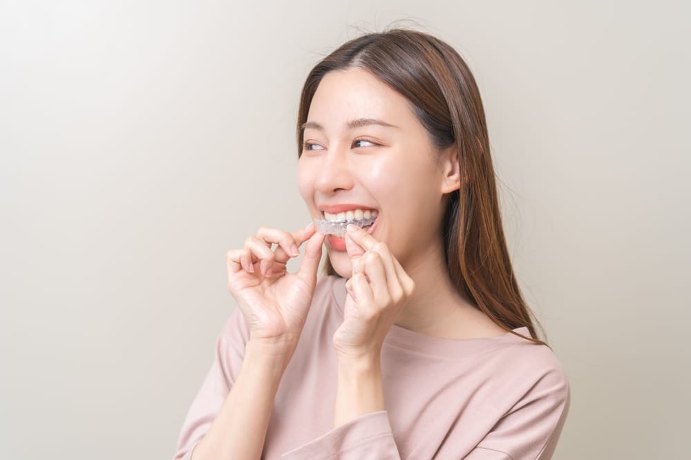 A woman smiles while holding a clear aligner near her teeth, preparing to place it in her mouth - Washington Orthodontics A woman smiles while holding a clear aligner near her teeth, preparing to place it in her mouth - Washington Orthodontics