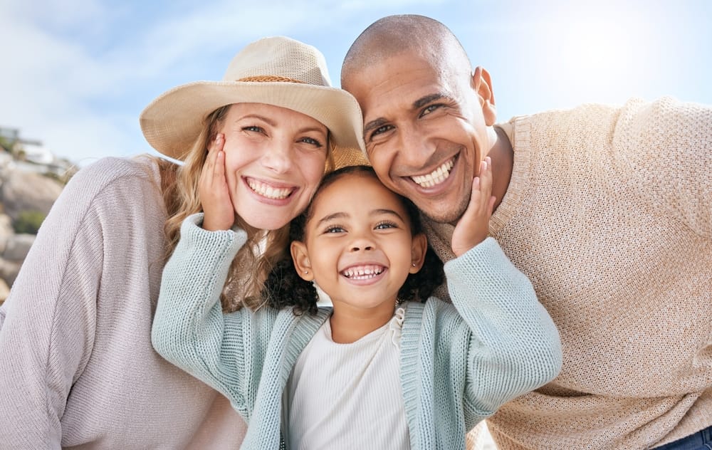 A mom, dad, and little girl, all smiling outdoors on a sunny day